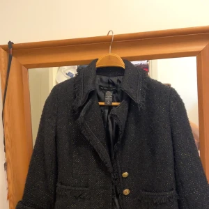 Svart kavajjacka med guldknappar från Zara - Säljer en svart kavajjacka från Zara med glittriga trådar och råa kanter. Jackan har tre dekorativa guldknappar framtill och två fickor. Perfekt för dig som vill ha en stilren men ändå unik look.