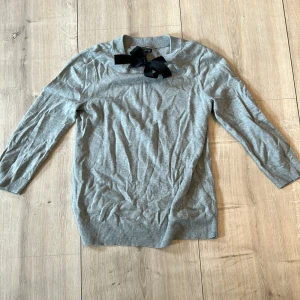 Grå tröja med svart rosett från Lindex - Säljer en ljusgrå tröja från Lindex med lång ärm och en dekorativ svart rosett vid halsen. Tröjan är i mjukt material och har en klassisk, rak passform. Perfekt för en stilren och söt look.