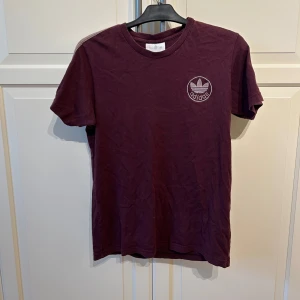 Vinröd vintage t-shirt från Adidas - Vinröd vintage t-shirt från Adidas med klassisk logga på bröstet och ljusa ränder längs ärmarna. T-shirten har rund halsringning och är tillverkad i mjuk bomull. Perfekt för en sport mm
