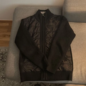 Svart jacka med stickade ärmar och quiltad front - Säljer en svart jacka med quiltad front och stickade ärmar. Jackan har dragkedja framtill och ribbade muddar. Perfekt för kyliga dagar när du vill ha både stil och komfort.