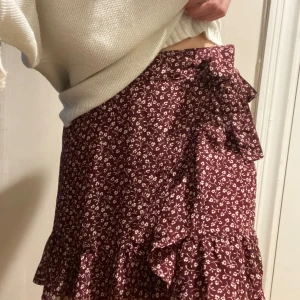 Vinröd blommig omlottkjol med volang - Supersöt vinröd omlottkjol med små vita blommor och volangkant. Kjolen har en knytning i midjan. Passar både till stickad tröja och t-shirt.