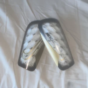 Nike Mercurial Lite benskydd - Säljer ett par Nike Mercurial Lite benskydd i svart, vitt och silver med hexagonmönster och gul detalj. Kommer med originalförvaringspåse. Lätta och smidiga för fotbollsträning eller match.