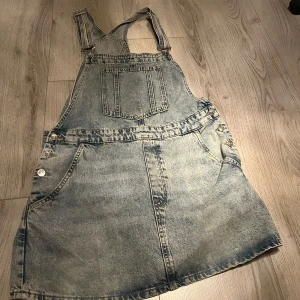 Jeansklänning med hängselband - Säljer en klassisk jeansklänning med hängselband och fickor fram och bak. Klänningen har knappar i sidorna och en stor bröstficka. Perfekt för en avslappnad och trendig look. Jeansklänningen är i väldigt bra skick och alla knappar är kvar. Hängslena gå att ändra.