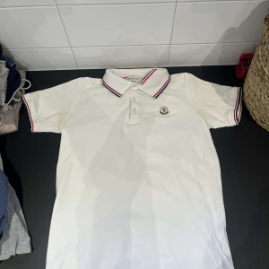 Vit pikétröja från Moncler - Säljer en klassisk vit pikétröja från Moncler med broderad logga på bröstet. Tröjan har krage med röd och blå rand samt knappar framtill. Perfekt för en stilren och sportig look.