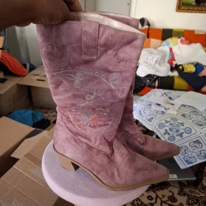Rosa mocka cowboy boots med broderi - Säljer ett par snygga rosa cowboy boots i mocka med broderade detaljer i vitt och pastellfärger. Skorna har en bred klack och rund tå, samt dekorativa sömmar och mönster längs skaftet. Perfekta för dig som vill sticka ut med din stil!