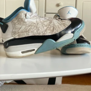 Nike Air Jordans med blå detaljer - Säljer ett par Nike Air Jordans med vit bas, svarta och ljusblå detaljer samt unikt mönster på ovandelen. Skorna har snörning, rund tå och platt sula. Perfekta för dig som gillar sneakers med lite extra stil.