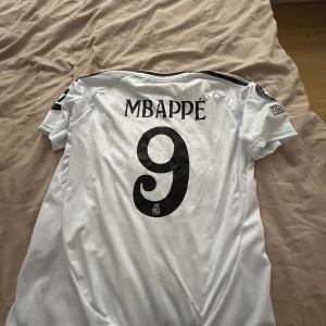 Real Madrid Mbappé 9 matchtröja - Säljer en vit Real Madrid fotbollströja med Mbappé och nummer 9 på ryggen. Tröjan har svarta detaljer, tre ränder på axlarna, klubbmärke på bröstet och flera tryck på ärmarna. Adidas slim fit-modell. Priset kan diskuters 