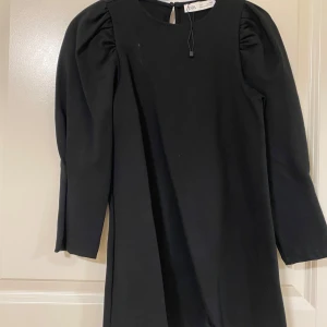 Svart klänning med puffärm från H&M - En svart klänning med rund halsringning och markerade puffärmar. Klänningen har trekvartsärm och en rak, enkel siluett som ger en stilren look. Perfekt för dig som gillar minimalistisk stil med en twist.