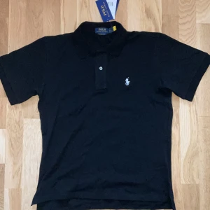 Svart pikétröja från Polo Ralph Lauren - Klassisk svart pikétröja från Polo Ralph Lauren med vit broderad logga på bröstet. Tröjan har korta ärmar, krage och knäppning med två knappar. Perfekt för en stilren och tidlös look.