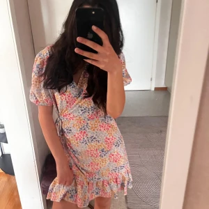 Blommig omlottklänning från Gina Tricot - Söt kort blommig omlottklänning från Gina Tricot med volangdetaljer och puffärmar. Klänningen har knyt i midjan och v-ringning. Perfekt för sommarens alla tillfällen!