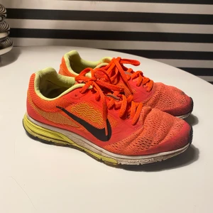 Nike löparskor i orange och gul - Nike löparskor i en snygg orange färg med gula detaljer och svart swoosh. Skorna har snörning och meshmaterial för bra andningsförmåga. Sulan är vit och gul med Nike Zoom-teknologi för extra komfort vid löpning.