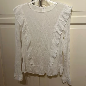 Vit blus med volangdetaljer från KappAhl - Säljer en vit långärmad blus från KappAhl i 100% bomull. Blusen har volangdetaljer framtill och baktill samt rund halsringning och dragkedja i ryggen. I strl S