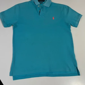 Turkos pikétröja från Polo Ralph Lauren - Säljer en turkos pikétröja från Polo Ralph Lauren med klassisk krage och knappslå. Tröjan har korta ärmar och den ikoniska röda logotypen broderad på bröstet. Slim fit-modell som passar perfekt till sommaren.