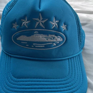 Blå keps med broderade stjärnor och båt - Snygg blå keps från Corteiz med vit brodyr framtill av stjärnor och en båt, samt fler stjärnor på sidan. Baksidan är i mesh och har justerbar knäppning. Perfekt för en cool och avslappnad stil.