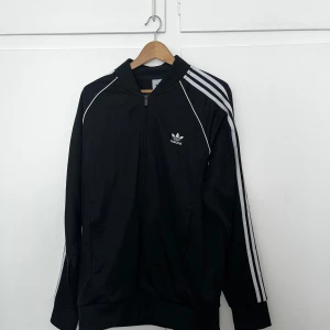 Svart Adidas track jacket med vita ränder - Jag säljer en svart Adidas SST träningsjacka med vita ränder längs ärmarna. Jackan har dragkedja på framsidan, två fickor och Adidas-logga på bröstet. Perfekt för en sportig och avslappnad stil. Nypris = 829kr, köpt på Zalando. Säljes för 300kr. Använd ett fåtal gånger.