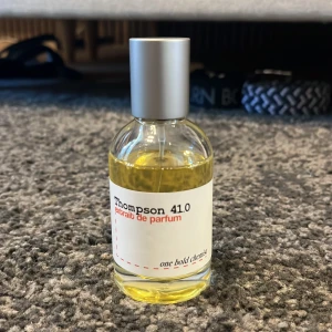 Thompson 41.0 Extrait de Parfum - En stilren parfymflaska med silverfärgad kork och transparent glas som visar den gula vätskan. Etiketten är vit med svart och röd text där det står 'Thompson 41.0 extrait de parfum' och 'one bold choice'. Perfekt för dig som gillar unika dofter. Inspirerad av Tom Ford Tobacco Vanille luktar som en söt Tobacco Honung med Vanilj riktigt god 45/50 Ml kvar