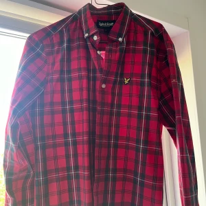 Rutig röd skjorta från Lyle & Scott - Säljer en klassisk röd och mörkgrön rutig skjorta från Lyle & Scott med button-down krage och broderad logga på bröstet. Skjortan har långa ärmar och knäppning framtill. Perfekt för dig som gillar stilrena mönster!