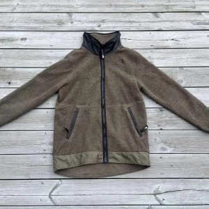 Olivgrön fleecejacka från The North Face - Nästintill oanvänd olivgrön fleecejacka från The North Face med svarta detaljer och hög krage. Jackan har dragkedja framtill, två fickor med dragkedja och förstärkt nederkant. 