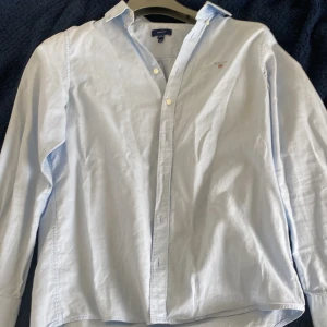 Ljusblå skjorta från GANT - Klassisk ljusblå skjorta från GANT med knappar framtill och krage. Skjortan har lång ärm och en liten broderad logga på bröstet. Perfekt för en stilren och tidlös look.