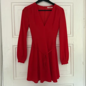 Röd omlottklänning från Glamorous - Säljer en röd omlottklänning från Glamorous med v-ringning och långa ärmar. Klänningen har knyt i midjan och en lätt utställd kjol. Perfekt för dig som vill ha en feminin och stilren look. 