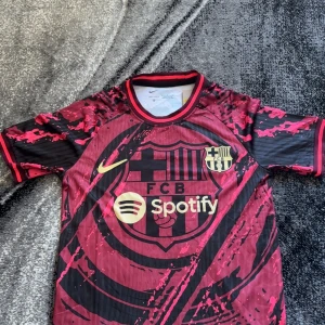 FC Barcelona x Spotify matchtröja - Säljer en FC Barcelona fotbollströja i röd och svart med gult Nike-märke och Spotify-logga på bröstet. Tröjan har korta ärmar och ett unikt mönster över hela tyget. Perfekt för dig som älskar Barca!
