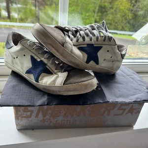 Golden Goose sneakers med blå stjärna - Säljer ett par vita Golden Goose sneakers med blå stjärna på sidan och grå snörning. Dom är i okej skic. Lite skavanker och dom är smutsiga men är fullt fungerande. Har låda och dustbag.