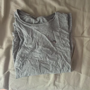 Grå t-shirt från GANT med logga - Snygg grå t-shirt från GANT med liten broderad logga på bröstet. Klassisk rund hals och korta ärmar, perfekt för en chill och enkel stil. Mjuk bomull som känns skön mot huden. Passar dig som gillar stilrena och tidlösa plagg.