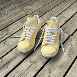 Adidas Stan Smith ljusgula sneakers - Fräscha Adidas Stan Smith sneakers i en mjuk ljusgul färg med vita detaljer och snörning. Skorna har en klassisk rund tå och platt sula, tillverkade i skinn med vit logga på hälen och tungan. Perfekt för dig som gillar stilrena och färgglada sneakers.