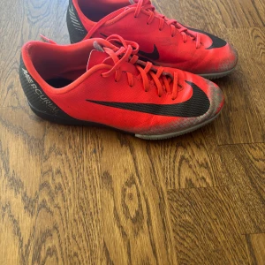 Nike Mercurial röda fotbollsskor - Säljer ett par Nike Mercurial fotbollsskor i en riktigt snygg röd färg med svarta detaljer och logga. Skorna har snörning och en platt sula med mönstrad undersida för bra grepp. Perfekta för dig som vill sticka ut på planen och ha maxad kontroll. Ingen låda tyvärr