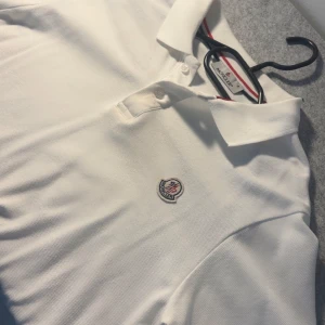 Vit Moncler piké  - Skicka dm för video/prisförslag! Storlek ”S” passar även M. Vit pikétröja från Moncler med klassisk krage och korta ärmar. Tröjan har två knappar vid halsen och Monclers ikoniska logga broderad på bröstet. Materialet är mjuk bomull och insidan av kragen har en snygg röd och blå rand.