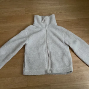 Vit teddyjacka med dragkedja, S - Mysig vit teddyjacka i storlek S med hög krage och hel dragkedja framtill. Jackan har två stora fickor och är tillverkad i fluffigt fleece-material som ger en skön och chill vibe. Perfekt för kalla dagar när du vill vara både bekväm och snygg. Hör av dig om du vill ha det billigare så kollar vi på det.