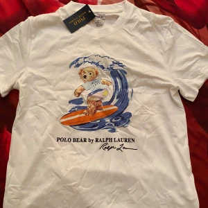 Vit Polo Bear t-shirt från Ralph Lauren - Vit t-shirt från Ralph Lauren med Polo Bear-tryck där nallen surfar på en orange bräda mot blå vågor. Rund hals och korta ärmar, tillverkad i mjuk bomull. Slim fit-modell som sitter snyggt och har en clean vibe med coolt motiv framtill.