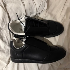 Svarta sneakers från Maison Margiela - Säljer ett par svarta sneakers från Maison Margiela med chunky siluett och bred plös. Skorna har svart snörning och kontrasterande vit insida. Tillverkade i slätt skinn med platt sula och rund tå. Perfekt för dig som gillar minimalistisk och edgy stil.
