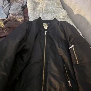 Svart bomberjacka från Saint Tropez - Svart bomberjacka från Saint Tropez med klassisk ribbad krage och ärmslut. Jackan har silverfärgade dragkedjor framtill och på ärmen, samt flera fickor. Materialet är blankt och känns lätt, perfekt för en cool streetstil. Storlek L