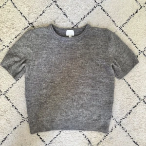 Grå stickad kortärmad tröja H&M - En enkel och snygg grå stickad tröja från H&M i storlek S. Modellen har korta ärmar, rund hals och ribbade muddar vid ärmslut och nederkant. Perfekt att matcha med jeans eller kjol för en chill look.