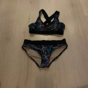 Blommig bikini med korsade band - Snygg bikini med blommigt mönster i blått, svart och röda toner. Toppen har breda korsade axelband och sportig form, medan trosan har bred svart kant upptill. Perfekt för dig som gillar färg!