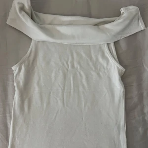 Vit offshoulder topp från Kappahl - Snygg vit offshoulder topp från Kappahl med bred krage som vilar över axlarna. Toppens stil är enkel och modern, perfekt för dig som gillar minimalistisk look. Materialet är mjukt och känns skönt mot huden. Passformen är normal och toppen är ärmlös.