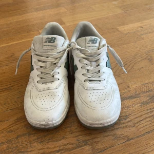 Vita och gröna New Balance sneakers - Säljer ett par fräscha New Balance sneakers i storlek 42. Skorna är vita med gröna detaljer på sidan och hälen, har rund tå och platt sula. Klassisk snörning och ventilerande perforeringar framtill. Perfekt för dig som gillar stilrena och sportiga sneakers.