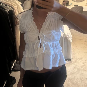 Vit volangblus med knyt - Supersöt vit blus med volanger och knyt framtill. Blusen har en v-ringad design, korta ärmar med rynkade detaljer och är croppad. 