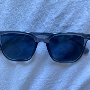 Blå transparenta solglasögon - Snygga solglasögon med transparent blå båge och mörkblå glas. Rektangulär form som ger en modern vibe och passar till de flesta ansikten. Lätta och bekväma att bära hela dagen. Perfekta för soliga dagar och en cool look.