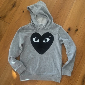Grå hoodie från Comme des Garçons Play - Säljer en grå hoodie från Comme des Garçons Play med det ikoniska svarta hjärtat med ögon på bröstet. Tröjan har huva med snörning, långärmade ärmar och en stor magficka. Perfekt för dig som gillar streetwear och vill sticka ut med en cool detalj.