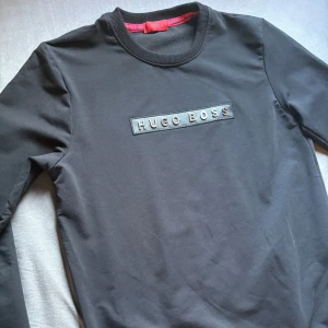 Svart sweatshirt från Hugo Boss - Svart långärmad sweatshirt från Hugo Boss med rund hals och ett stilrent broderat logotypmärke på bröstet. Tröjan har en klassisk passform och är gjord i mjuk bomull som känns skön mot huden. Perfekt för dig som gillar enkel och snygg design. Knappt använd, riktig fräsch