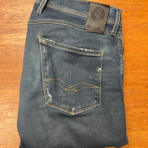 Replay Anbass Hyperflex - Snygga Replay Anbass Hyperflex jeans i klassisk blå färg med coola slitningar och kontrastsömmar på bakfickorna. Modellen har normal passform och raka ben, med stretchigt material som gör dem riktigt sköna. Perfekt för dig som vill ha stil och komfort i ett.