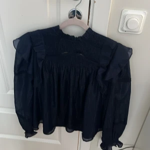 Mörkblå volangblus från Gina Tricot - Supersöt mörkblå blus från Gina Tricot med volangdetaljer på axlarna och lätt glansigt tyg. Blusen har lång ärm, rynkade partier upptill och liten ståkrage med volang. Perfekt för dig som gillar en romantisk och trendig look.