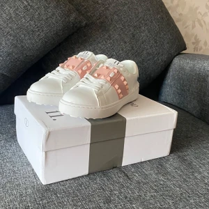 Valentino Rockstud sneakers vit/rosa - Säljer ett par Valentino Rockstud sneakers i vitt skinn med ljusrosa band och ikoniska nitar över ovansidan. Skorna har rund tå, platt sula och snörning. Märket syns på plösen och innersulan. Perfekt för dig som gillar edgy designerstil.                                                          ❗️Just Nu❗️20% rabatt när man följer mig!
