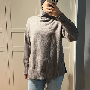 Grå polotröja med slits i sidan - Stickad polotröja från H&M med slits på sidorna och en liten slits i nacken. Färgen är en blandning av grått och beige. Materialet är mjukt och varmt. Fortfarande i bra skick men något nopprig och lite skrynklig efter tvätt.