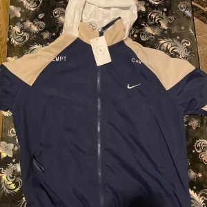 Nike x Cav Empt vindjacka blå/beige - Säljer en fet sällsynt vindjacka från Nike x Cav Empt i mörkblått och beige med vit huva. Jackan har broderade loggor, coolt grafiskt tryck på ryggen och dragkedja framtill. Materialet är lätt polyester, perfekt för streetwear. Snygga detaljer och clean design. Fick den som present men den var för stor för mig!!
