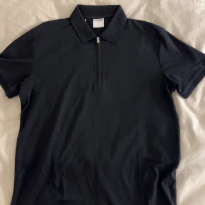 Svart pikétröja Selected Homme - Svart pikétröja från Selected Homme med klassisk krage och half zip framtill. T-shirten har korta ärmar och är gjord i mjuk bomull. Perfekt för en clean och stilren look, passar dig som gillar enkel design och diskreta detaljer.