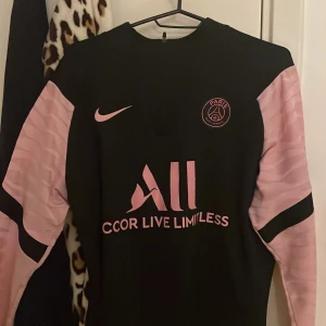 Svart och rosa PSG långärmad fotbollströja - Snygg PSG fotbollströja från Nike med svart kropp och ljusrosa långärmade ärmar med subtilt mönster. Tryck i rosa på bröstet och ryggen, inklusive Paris Saint-Germain-logga och sponsor. Materialet är lätt och andas, perfekt för träning eller match.
