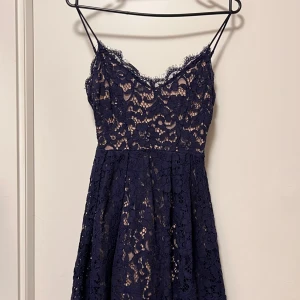 Marinblå spetsklänning med smala axelband - Säljer en marinblå klänning med smala axelband och vacker spets över hela klänningen. Den har en lätt utställd kjol och blommigt spetsmönster, samt nude-färgat foder under. Perfekt för dig som vill ha en elegant look med lite edge.